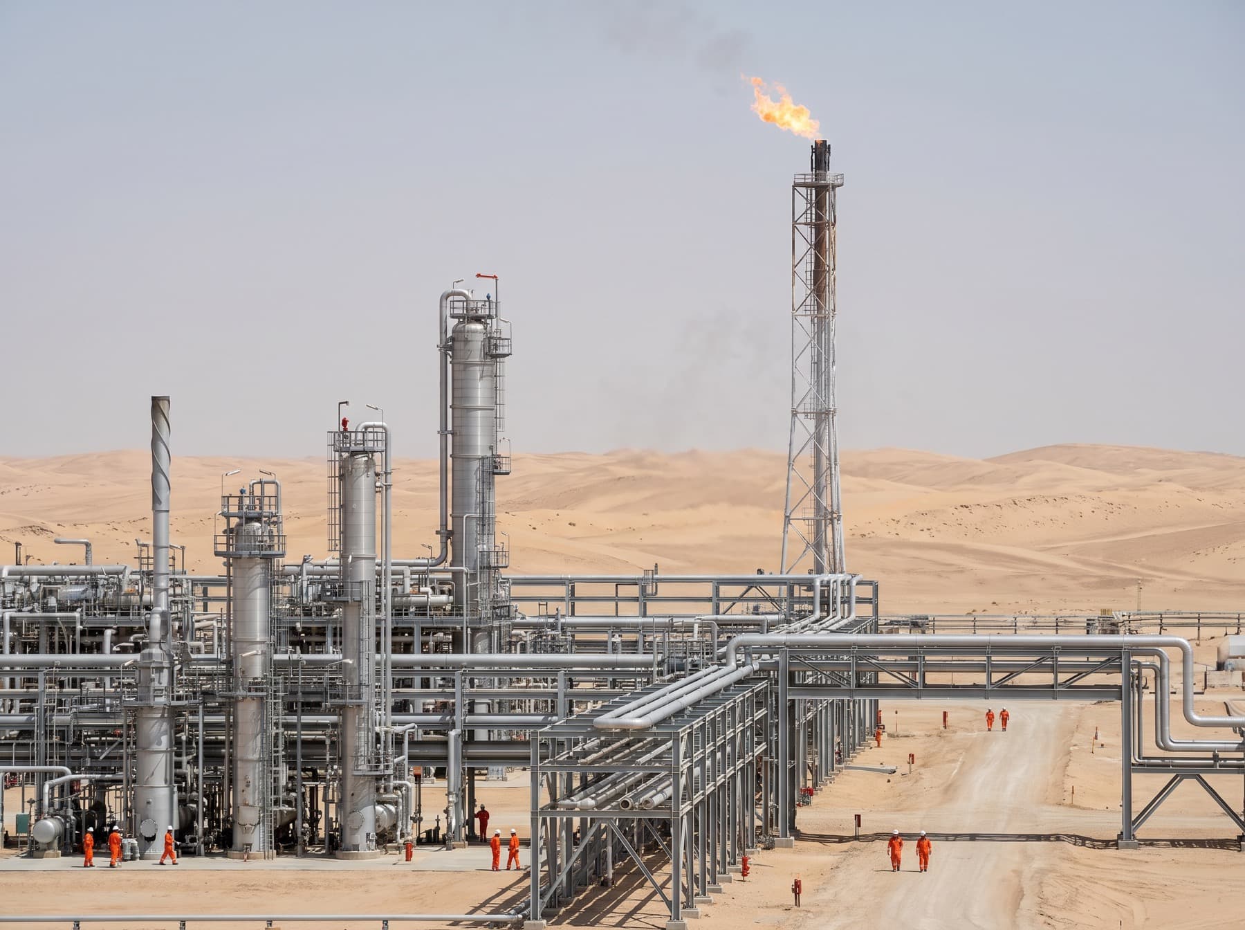 A gas processing plant in the desert at midday, a flare burning above the pipe racks.